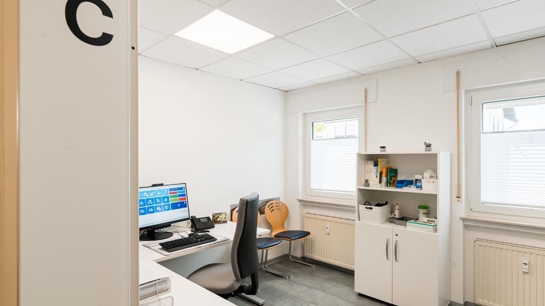 Hell eingerichtetes Sprechzimmer (Zimmer C) mit Schreibtisch, PC-Monitor, ergonomischem Bürostuhl und einem weißen Praxisschrank.